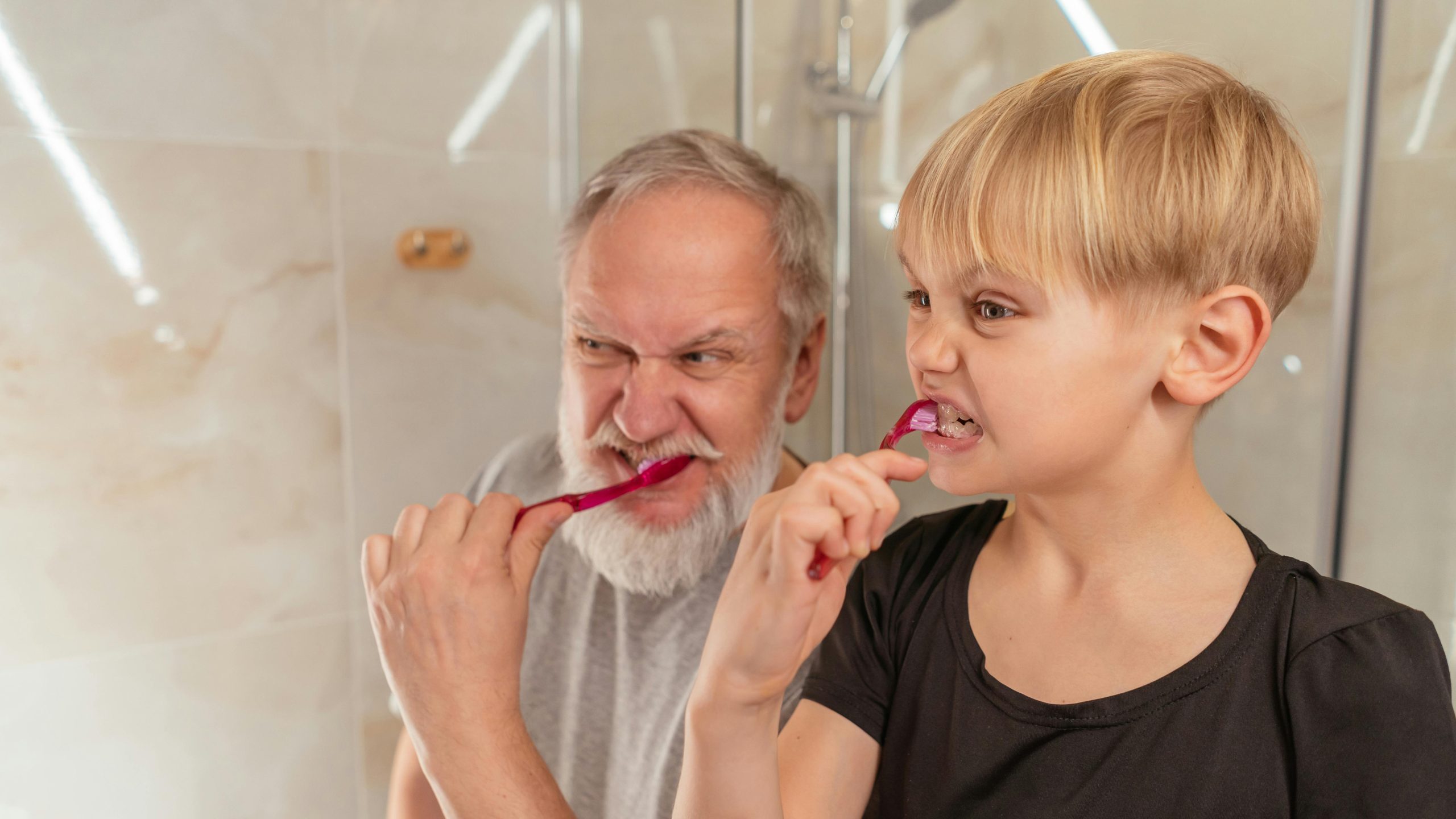 A young child and an older man stand in a bathroom brushing their teeth side by side, both making exaggerated, intense facial expressions while looking at a mirror.